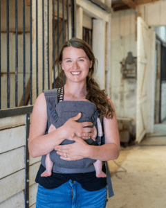 Kalen McKelvey Owner, Sunshine Stables is standing in a horse stable with a baby in a front carrier pack.